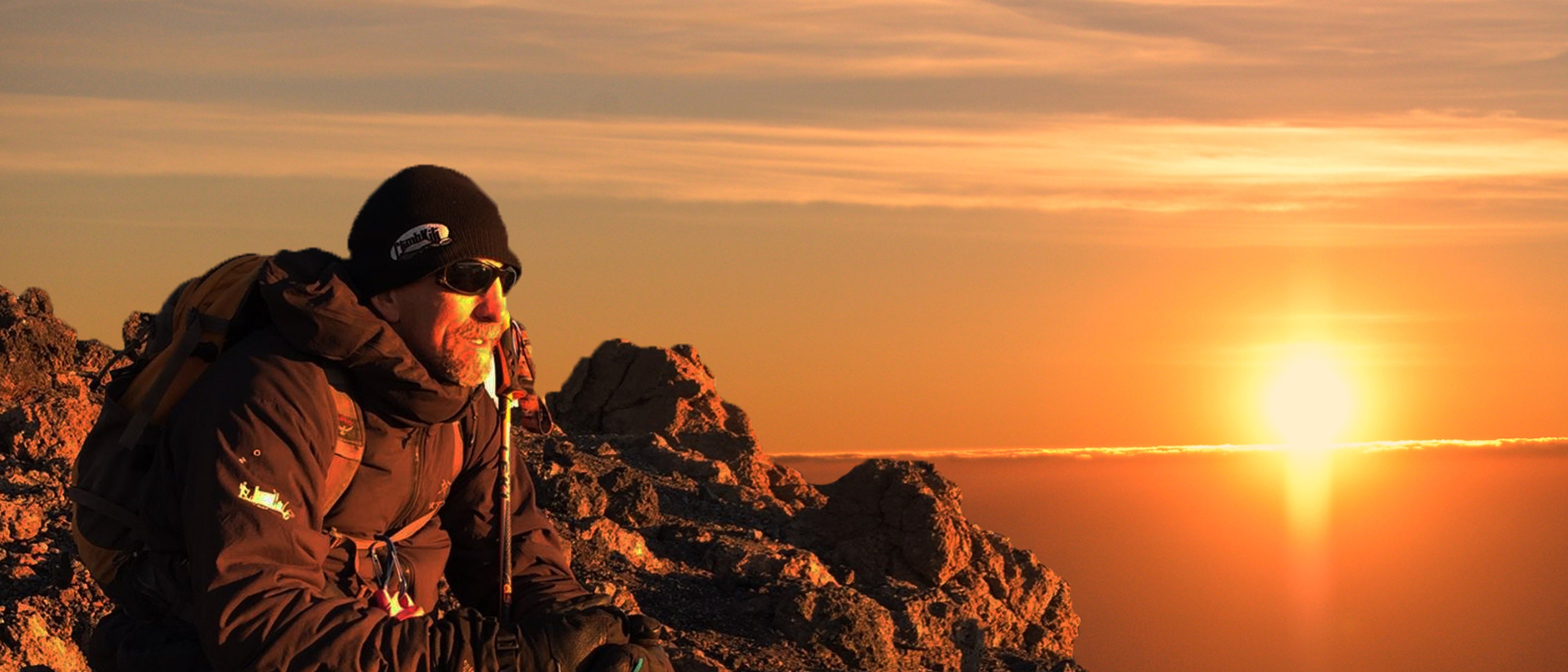 Randy Pierce, wearing dark climbing gear, sunglasses, a knit cap, and a large backpack, rests on a rugged rocky slope near the summit of Mount Kilimanjaro as the sun rises with dramatic colors and horizon.
